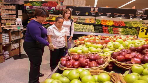 Three people stand in a grocery store produce section, examining baskets of red and green apples. Shelves display various colorful fruits and vegetables.