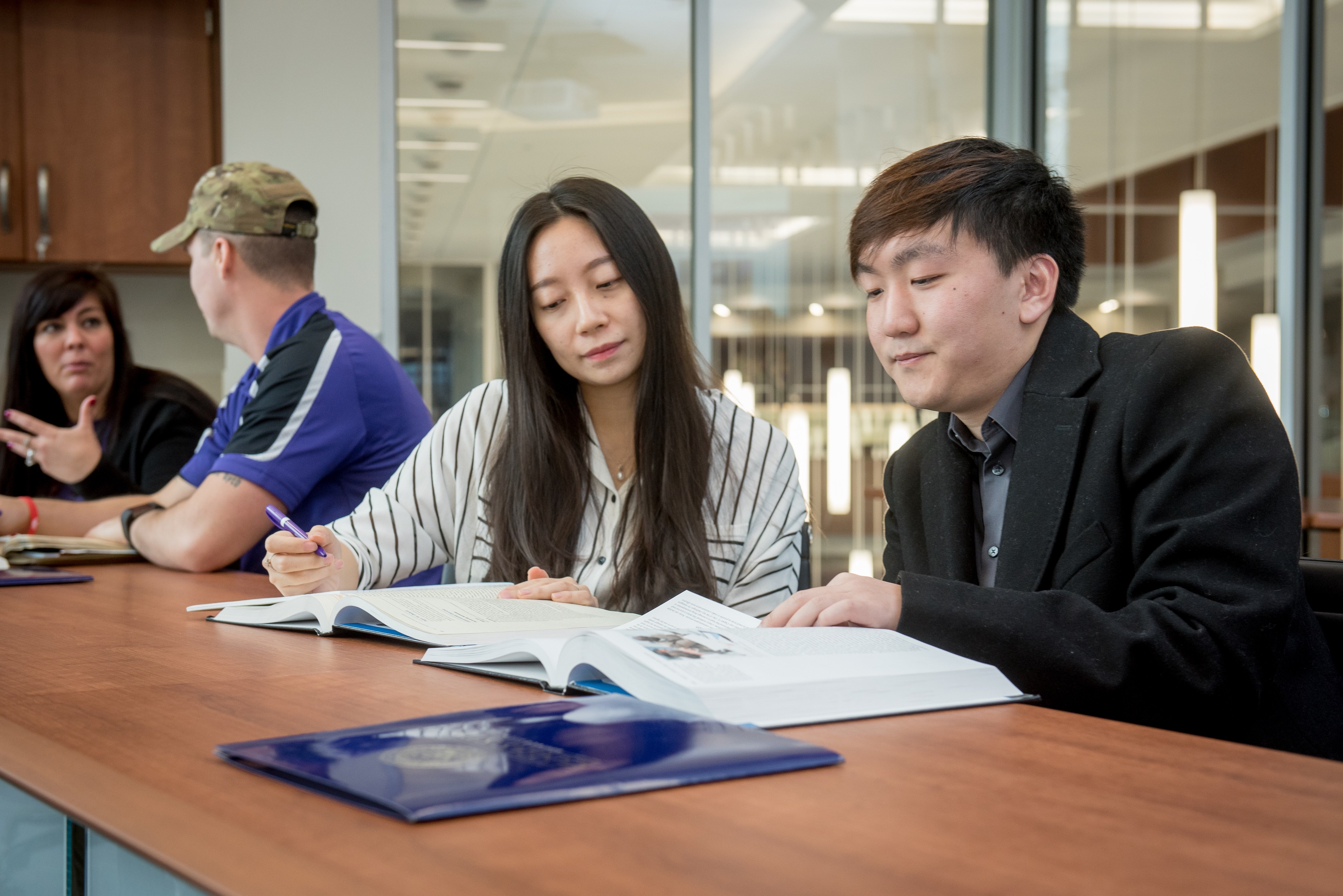 A group of four people sit at a table in a contemporary room. Two individuals in the foreground are focused on reading and discussing open textbooks, conveying a studious and collaborative atmosphere.
