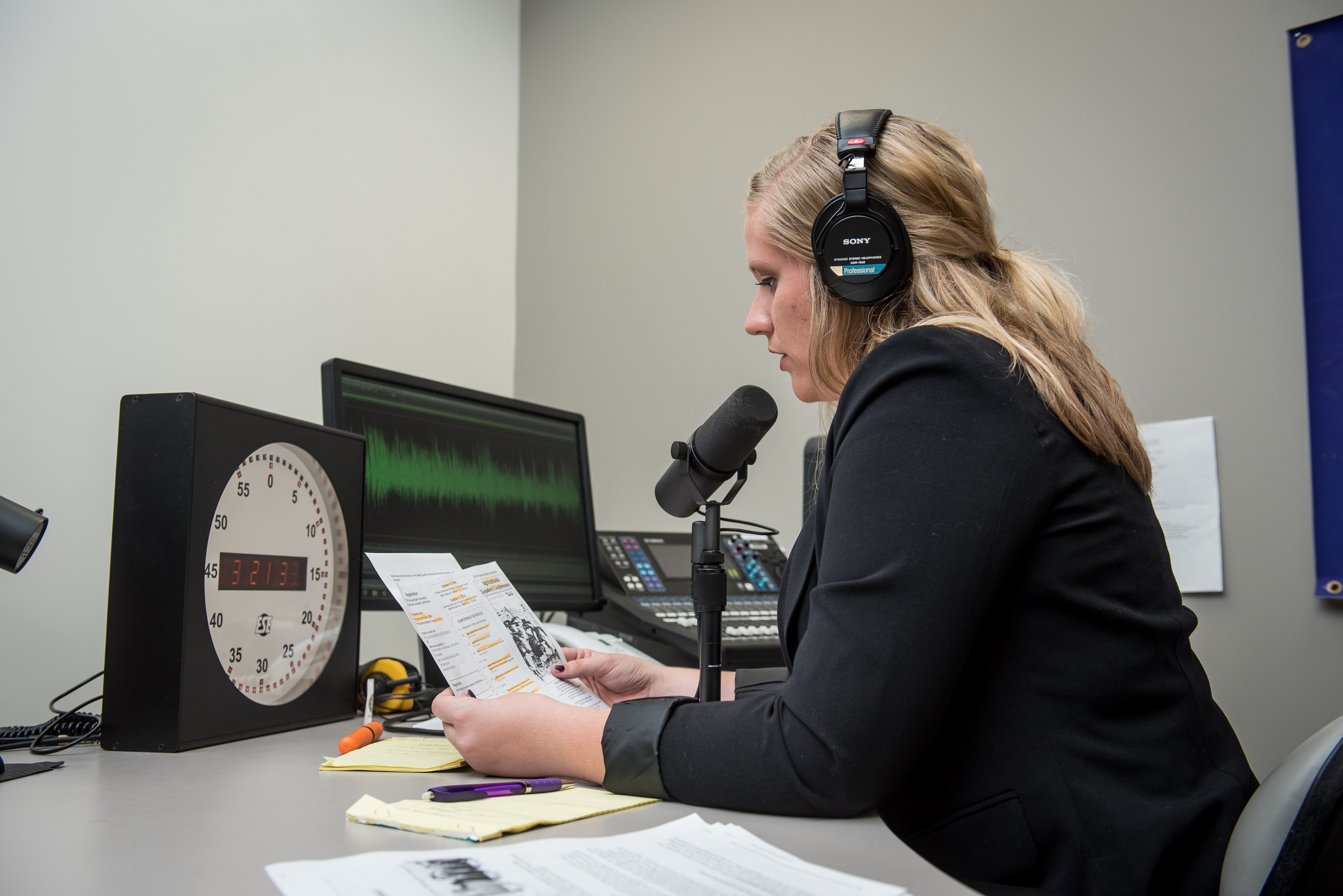A woman in headphones sits at a desk, speaking into a microphone in a recording studio. She holds papers, with a clock and audio monitor nearby.