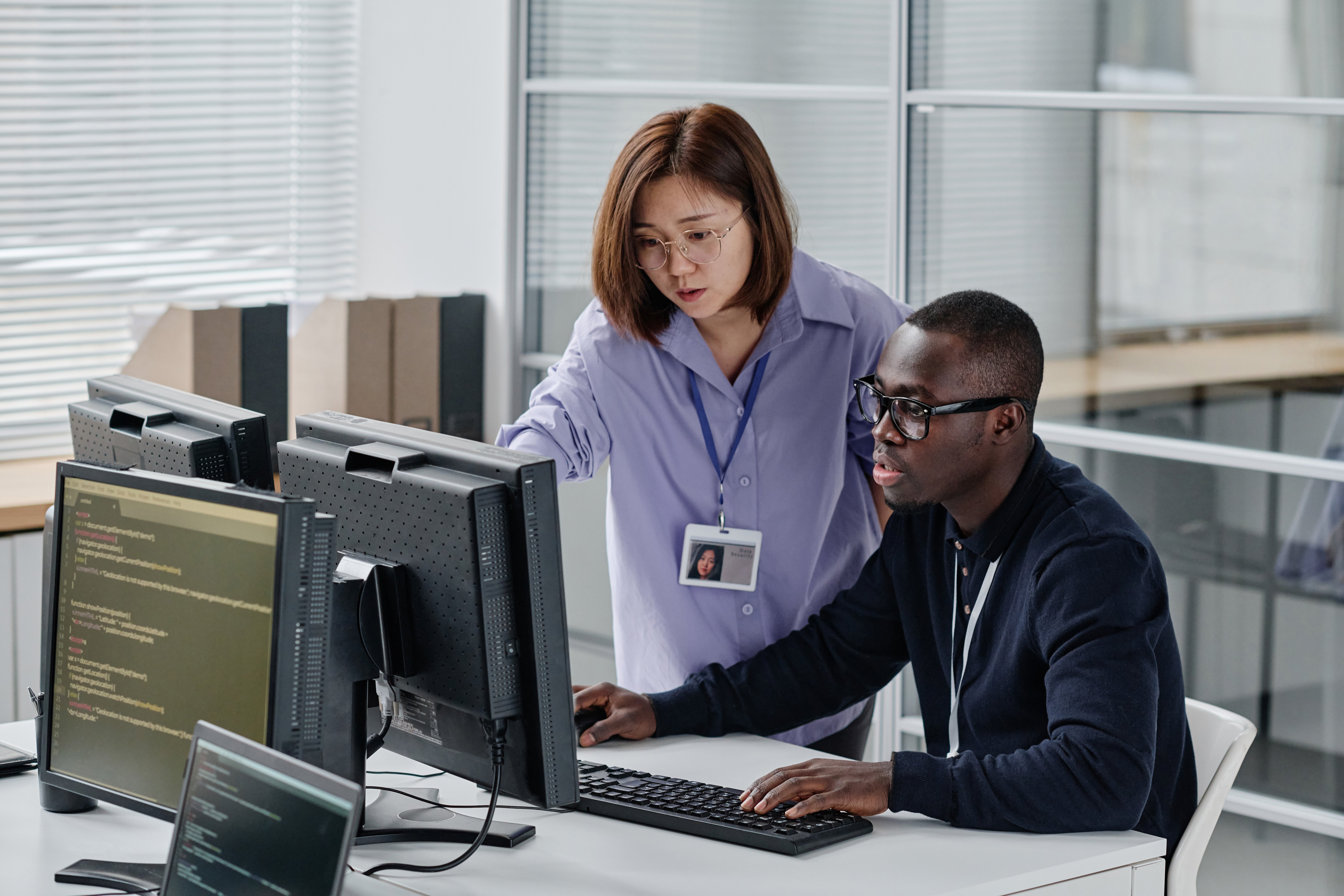 Two colleagues collaborate in an office. A woman points at a screen displaying code, assisting a man working intently on a computer. The atmosphere is focused.