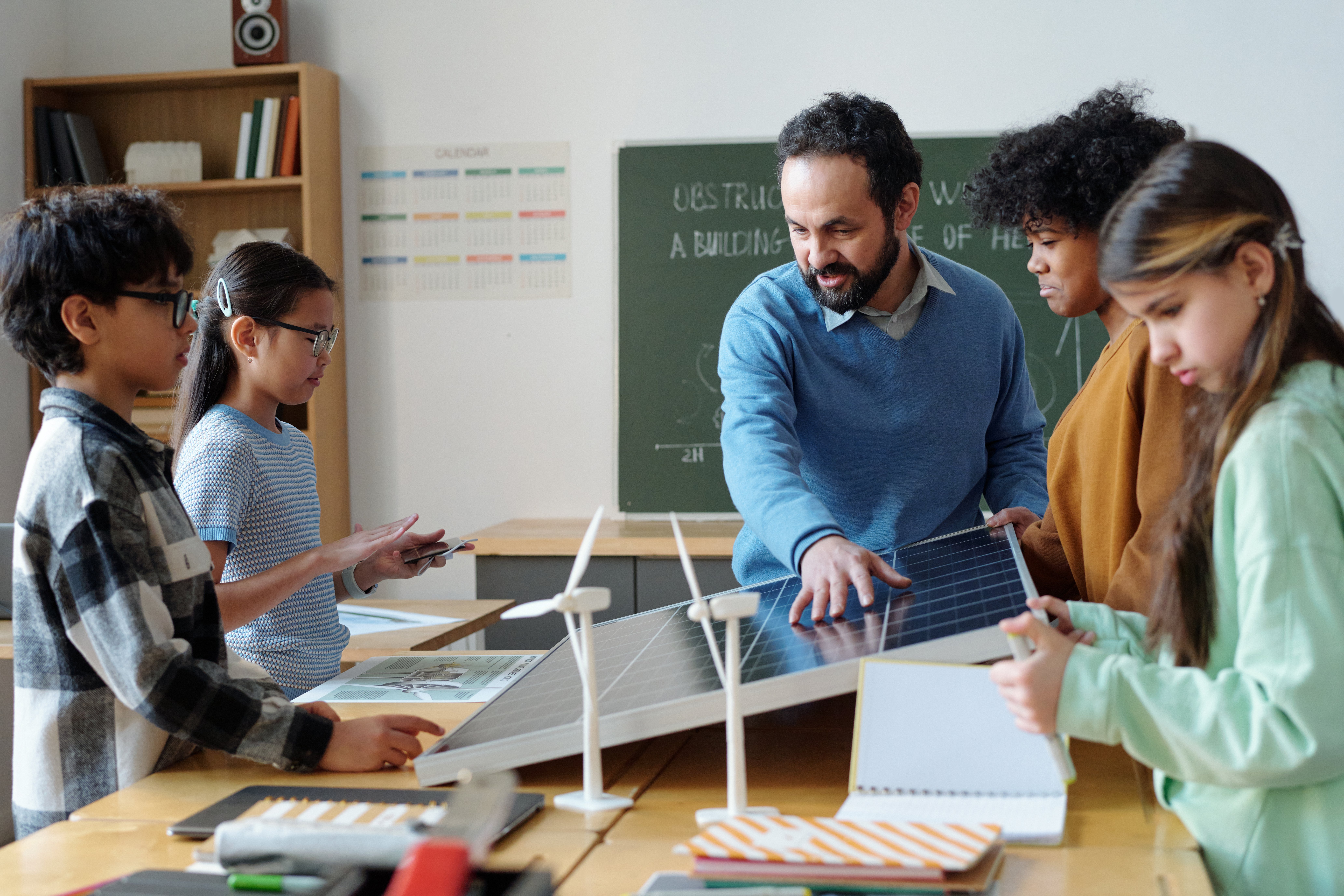A teacher and diverse group of students examine a solar panel and small wind turbines in a classroom, fostering curiosity and teamwork.