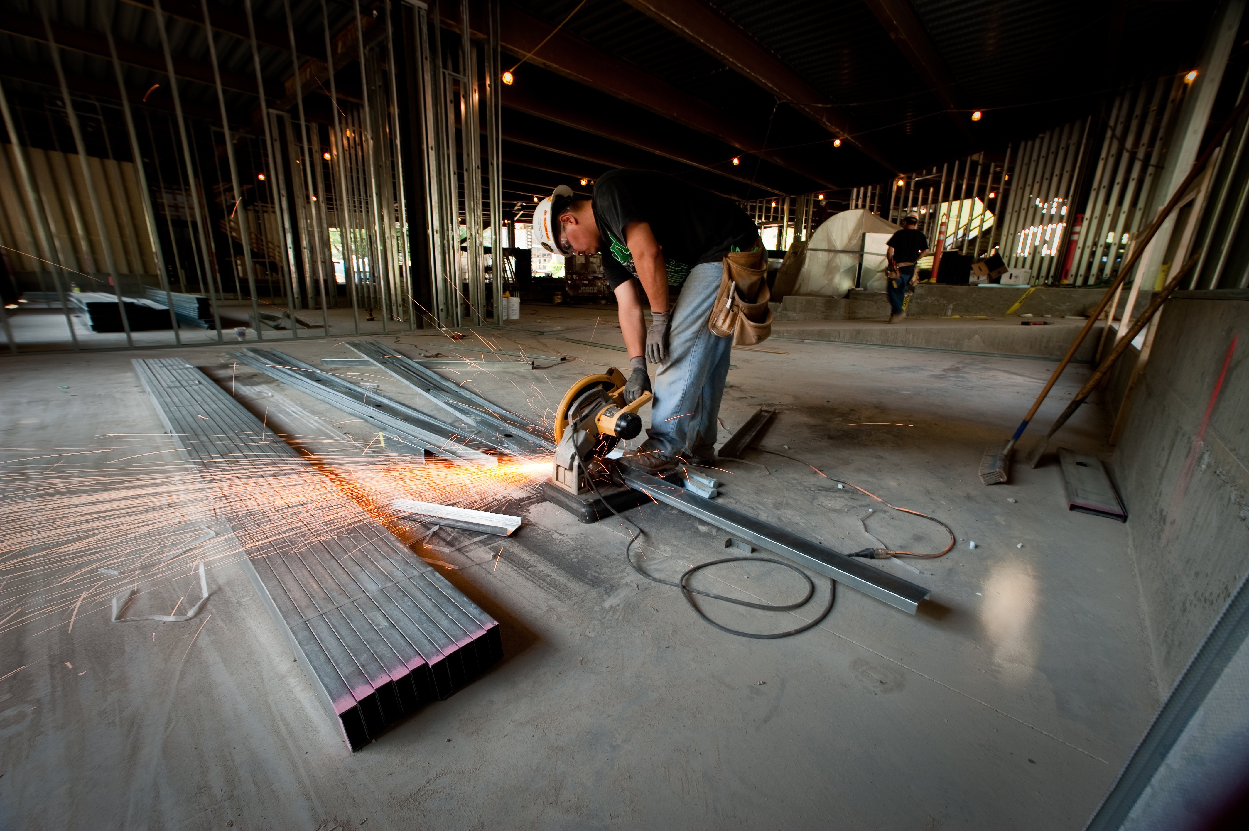 Worker using a power saw to cut metal beams in a dimly lit construction site. Sparks fly, creating a dynamic feel. Metal frames and tools surround him.