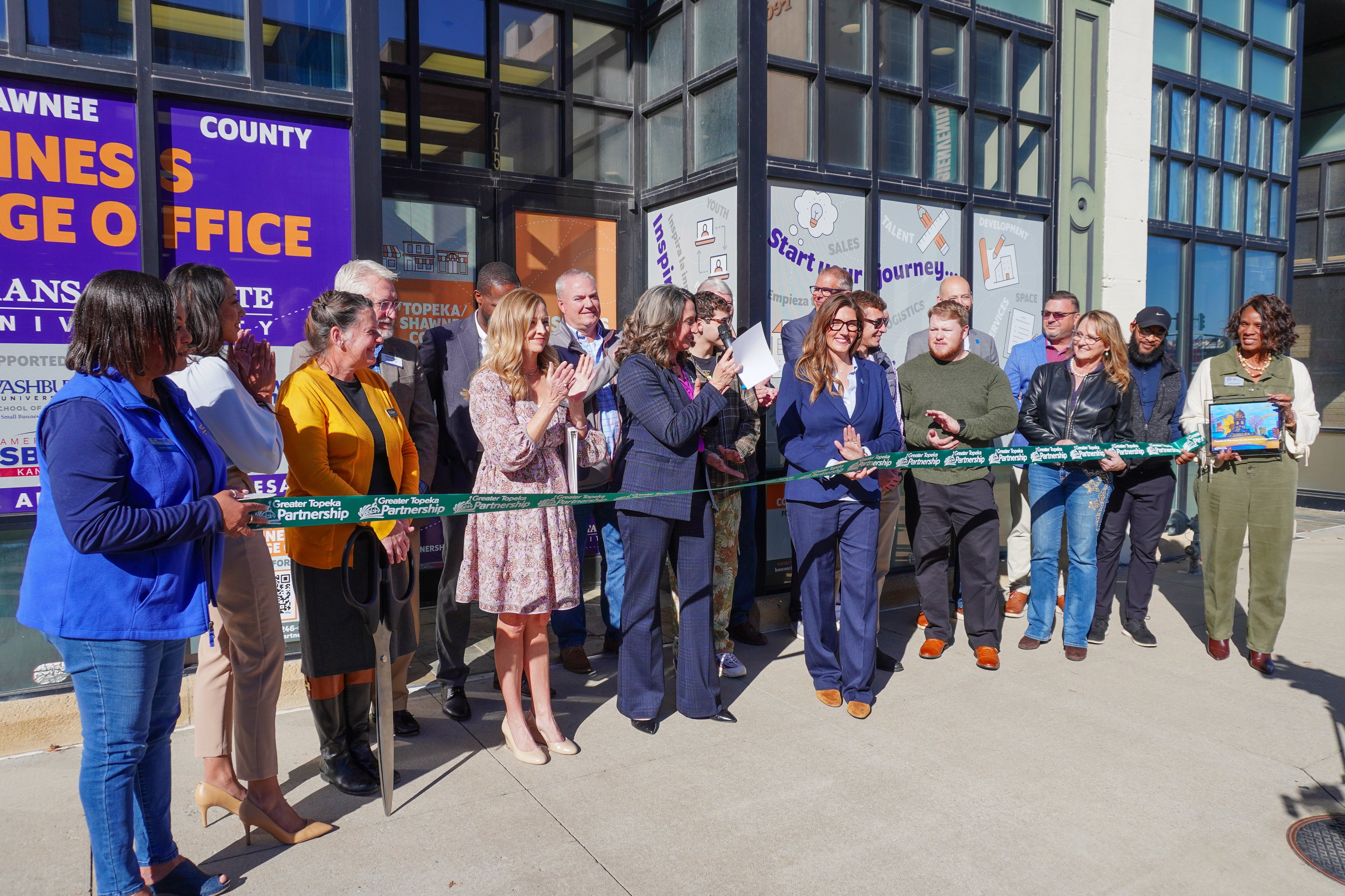 A group of people stand outside a building, holding a green ribbon for a ceremonial cutting. They are smiling, clapping, and appear celebratory.