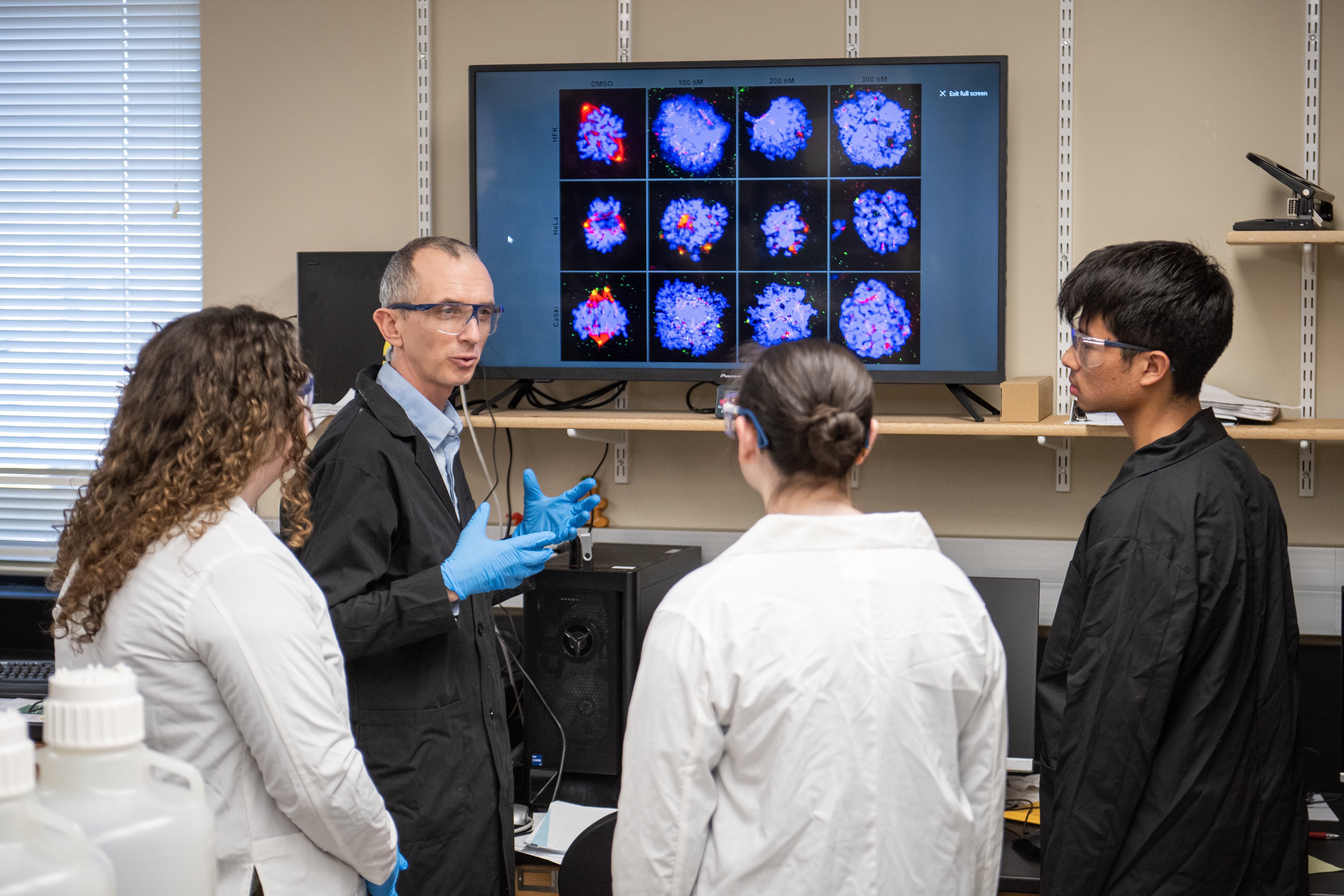 A scientist wearing gloves and speaking to three people in lab coats. They are viewing colorful scientific images on a screen in a laboratory setting.