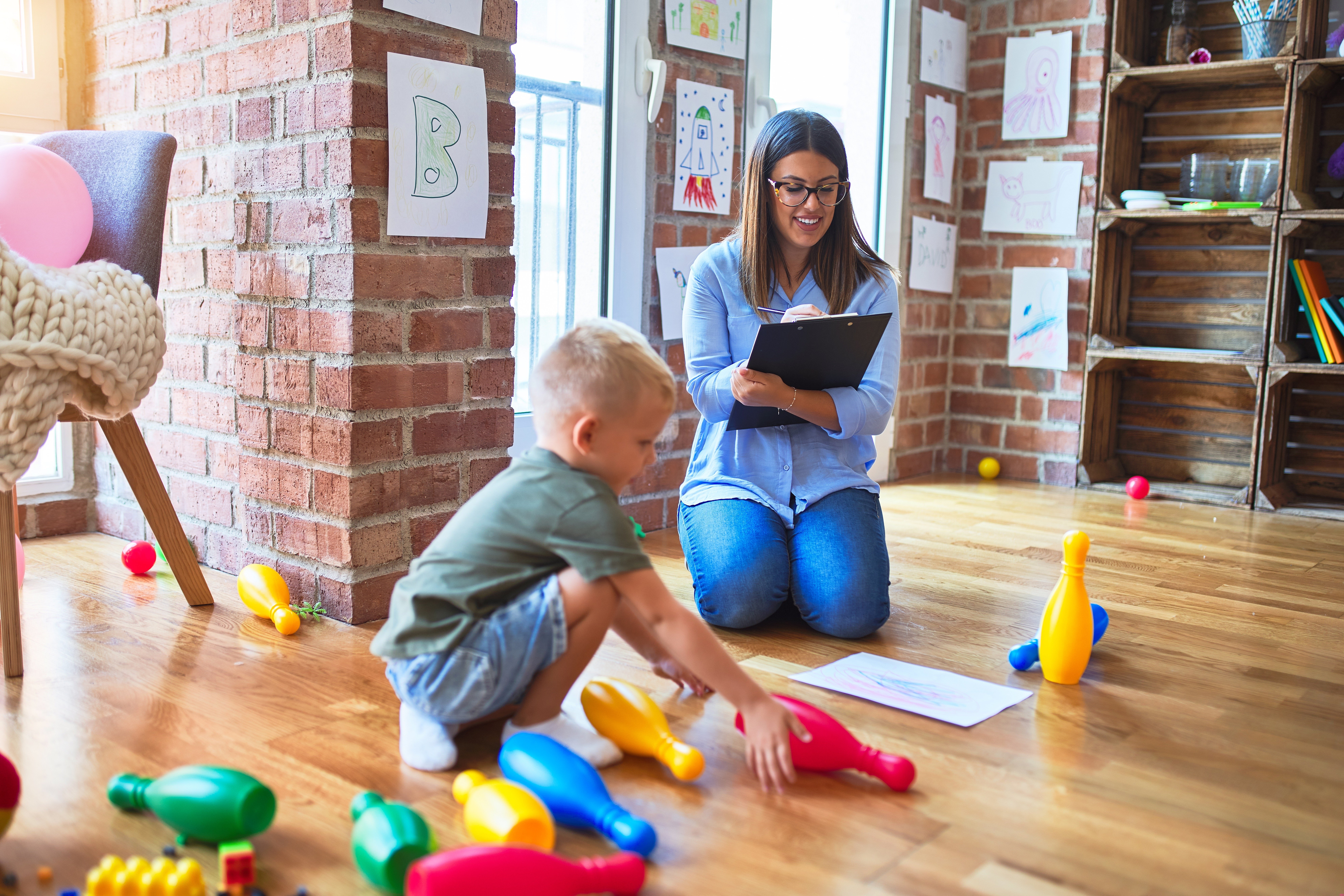 A young child plays with colorful toy bowling pins on a wooden floor, while a woman with glasses watches and takes notes on a clipboard. The room has brick walls and children's drawings displayed, creating a warm and playful atmosphere.
