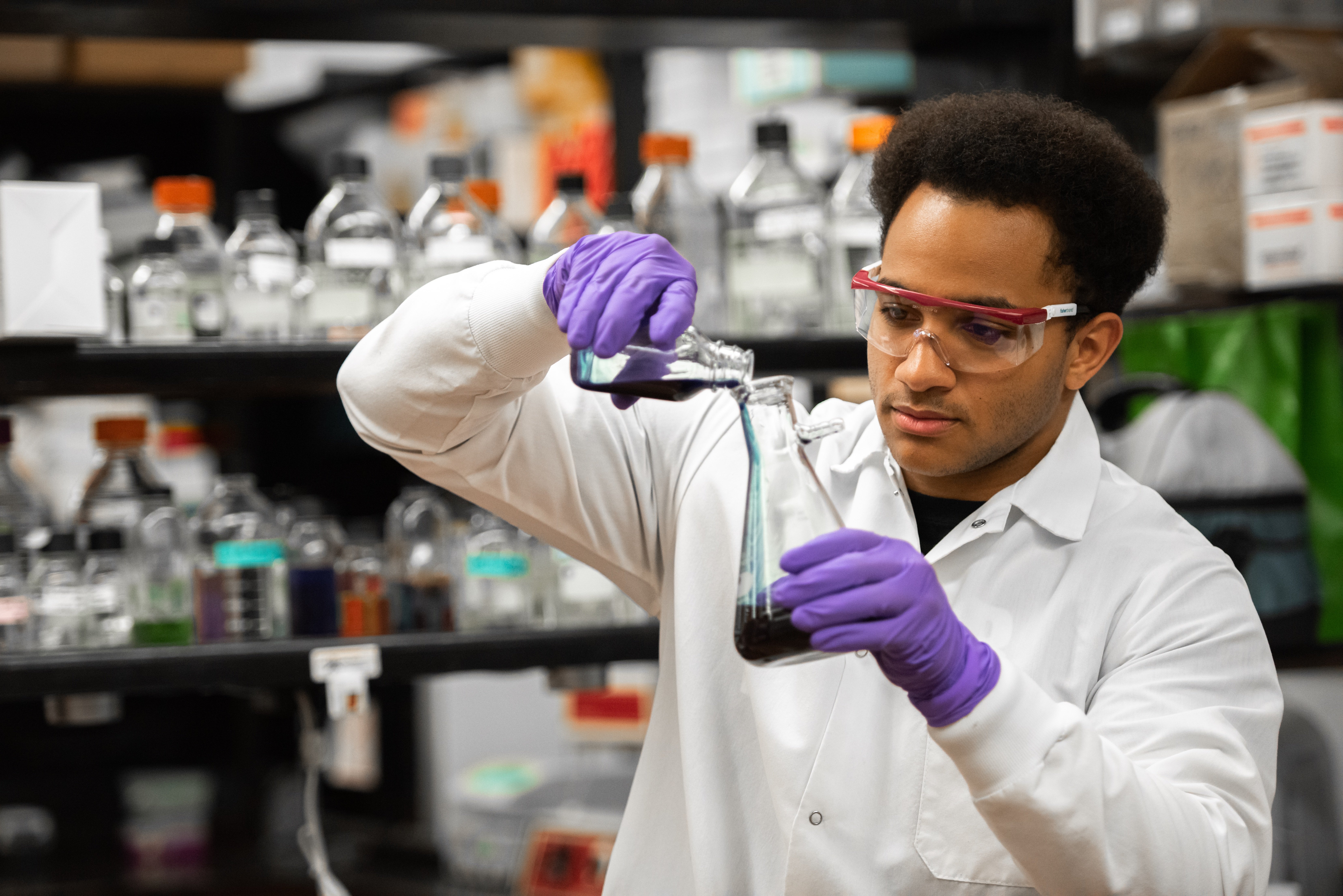 A scientist in a lab coat and safety goggles carefully pours liquid from one flask to another, surrounded by shelves of lab equipment. The scene conveys focus and precision.