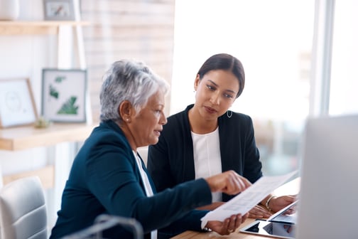 An older woman and a younger woman looking at paperwork at a desk in an office setting.